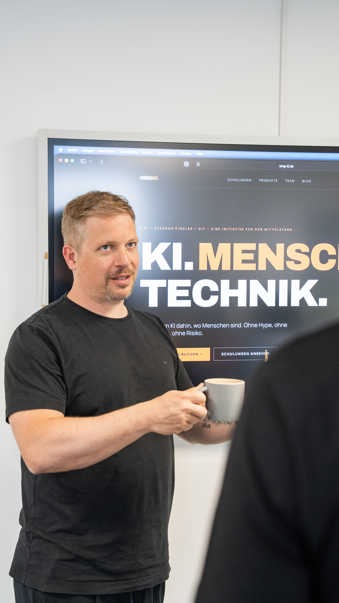 Andreas Olbricht, Geschäftsführer der OIT GmbH, im OIT-Büro in Osnabrück — mit Kaffee vor dem KI.MENSCH.TECHNIK.-Hero-Screen der OMG-KI-Initiative.